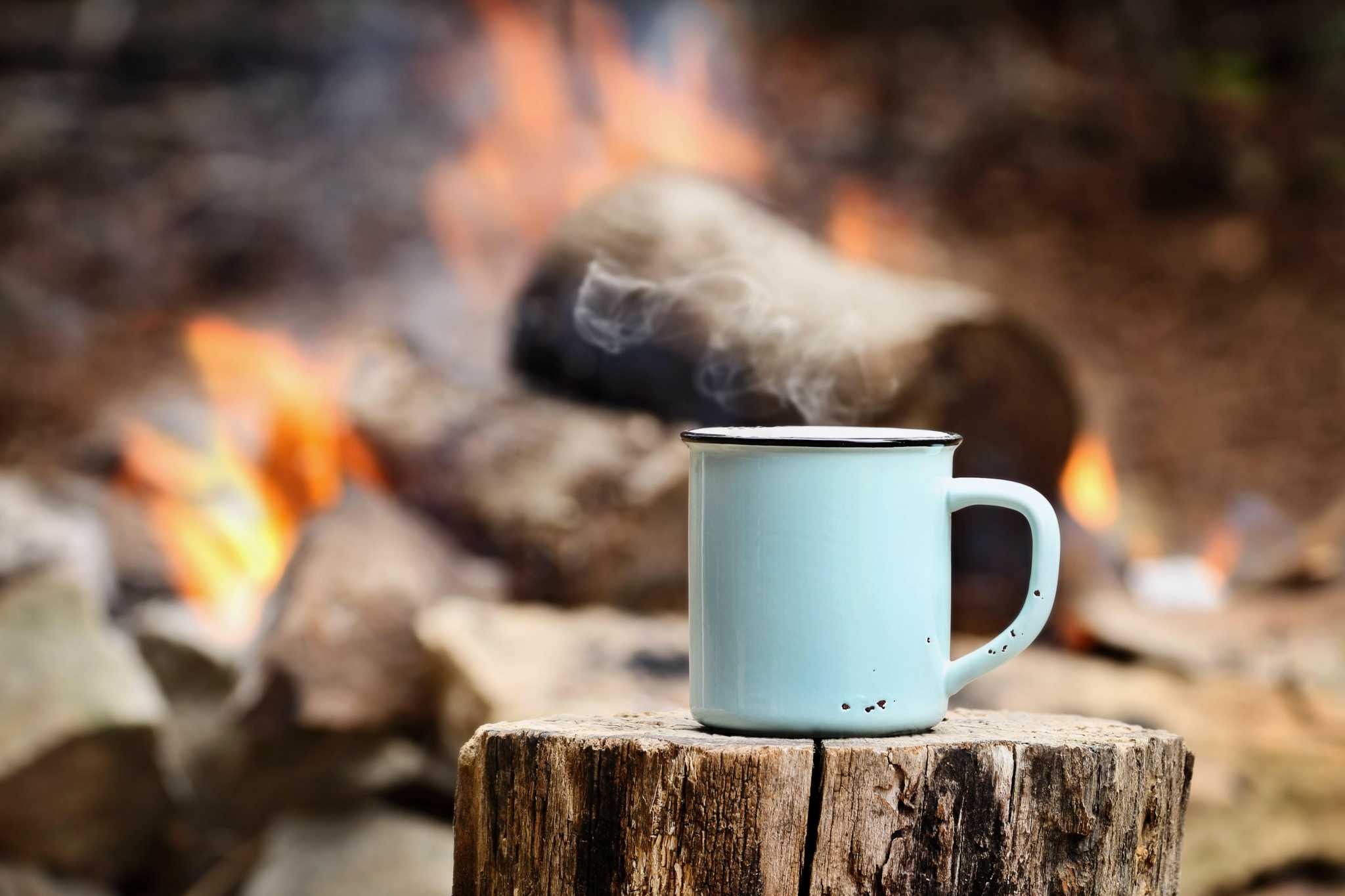 Metal mug with steaming coffee in front of a campfire.